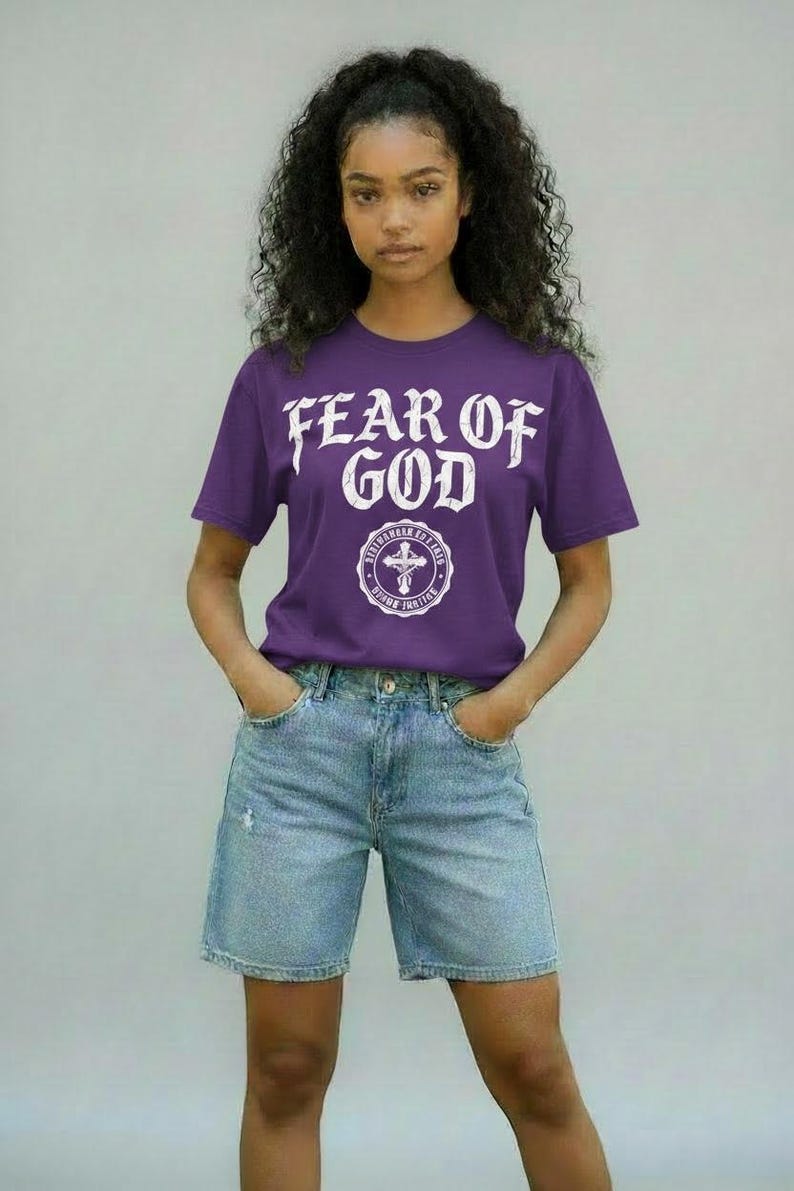 May include: A purple t-shirt with the text "FEAR OF GOD" in white, above a circular emblem with a cross. The model is wearing light blue denim shorts. The background is a solid light gray.