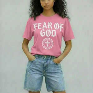 May include: Pink t-shirt with the text "FEAR OF GOD" in white, above a circular emblem with a cross. The shirt is paired with denim shorts. The overall style is casual and modern.