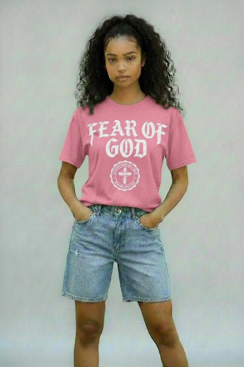 May include: Pink t-shirt with the text "FEAR OF GOD" in white, above a circular emblem with a cross. The shirt is paired with denim shorts. The overall style is casual and modern.