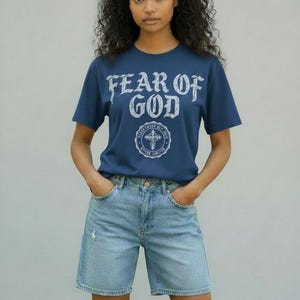 May include: A navy blue t-shirt with the text "FEAR OF GOD" in a distressed white font. Below the text is a circular emblem with a cross. The shirt is paired with light blue denim shorts.
