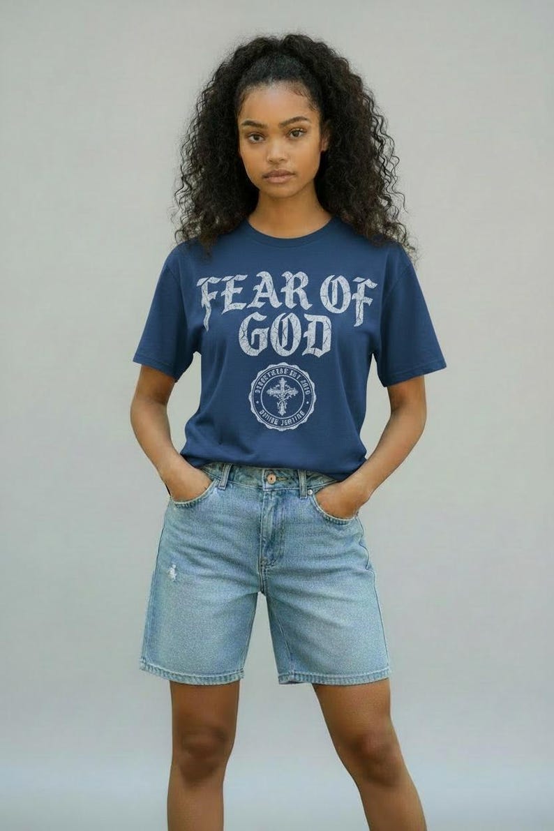 May include: A navy blue t-shirt with the text "FEAR OF GOD" in a distressed white font. Below the text is a circular emblem with a cross. The shirt is paired with light blue denim shorts.