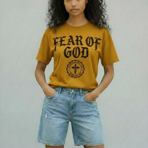 May include: Mustard yellow t-shirt with the text "FEAR OF GOD" in a distressed font. Below the text is a circular emblem with a cross. The shirt is paired with light blue denim shorts.