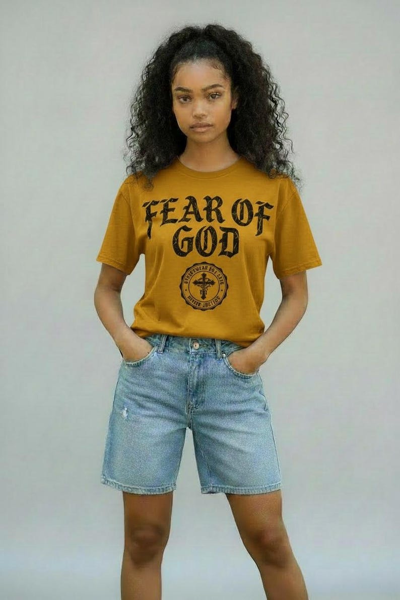 May include: Mustard yellow t-shirt with the text "FEAR OF GOD" in a distressed font. Below the text is a circular emblem with a cross. The shirt is paired with light blue denim shorts.