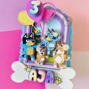 May include: A layered paper cake topper featuring cartoon dogs, balloons, and a number 3. The topper includes the name "AJA" and paw prints. The color scheme includes pink, blue, yellow, and white.