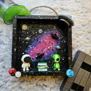 May include: A black, square, space-themed shadow box with a painted galaxy background. It features miniature figurines of an astronaut, an alien, UFOs, planets, and books. A green slime container and a gray, geometric structure are also present.