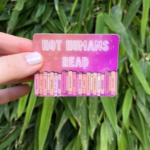 May include: A rectangular sticker with a pink and purple gradient background featuring the white text HOT HUMANS READ. Below the text is a row of illustrated book spines in shades of pink, each labeled with genres including Romance, Sci-Fi, Fantasy, Thriller, Young Adult, Historical Fiction, Biography, Poetry, Horror, Smut, Contemporary, Dystopian, Memoir, Dark Romance, and Mystery.