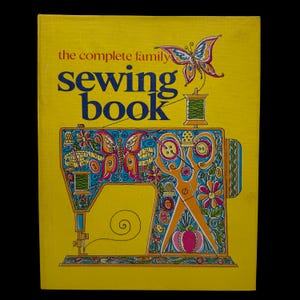May include: A bright yellow hardcover book titled the complete family sewing book. The cover features a large, colorful illustration of a sewing machine decorated with butterflies, flowers, buttons, and scissors, with a spool of thread above.