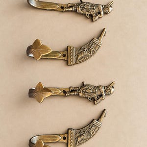 May include: Four antique brass-toned metal wall hooks arranged vertically. Two hooks feature a relief of a mounted archer, while the other two depict a figure in armour. Each hook ends in a three-leaf design and includes a small screw hole for mounting.
