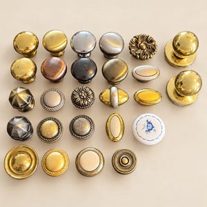 May include: A collection of twenty-three assorted cabinet knobs arranged in rows. The knobs feature various shapes, including round, oval, and floral designs, with finishes in polished brass, brushed nickel, antique bronze, and one white ceramic knob.