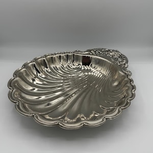 May include: A silver-coloured, shell-shaped serving dish with a scalloped edge. The dish has a textured, ridged interior and decorative floral detailing on one end. The dish is likely made of metal and is suitable for serving food or as a decorative item.