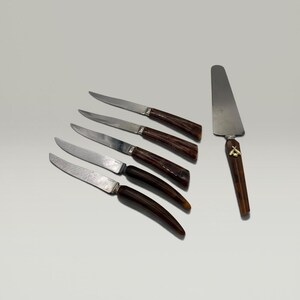 Könnte beinhalten: Ein Set aus fünf Steakmessern mit geschwungenen, dunkelbraunen Griffen und silberfarbenen Klingen, angeordnet in einer diagonalen Reihe, begleitet von einem flachen Tortenheber mit passendem dunkelbraunen Griff und einem kleinen goldenen Bienenemblem.