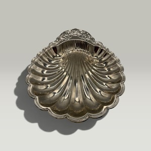 May include: A polished silver-toned metal dish shaped like a scallop shell. The dish features a scalloped edge and a series of radiating fluted grooves that extend from the base to the rim, with decorative embossed detailing at the top hinge.