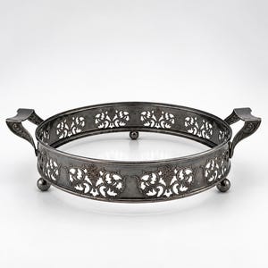 May include: A round silver-toned metal serving tray featuring an intricate openwork filigree pattern along the sides. The tray includes two decorative handles on opposite sides and is supported by three small, rounded ball-shaped feet.