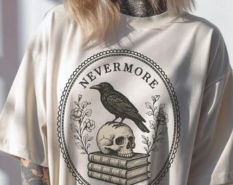 May include: Cream-colored t-shirt featuring a circular design with the word "NEVERMORE" in a decorative font. The design includes a black raven perched on a skull atop stacked books, surrounded by floral elements.