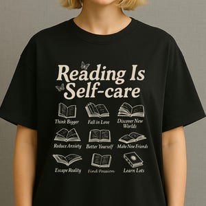 Puede incluir: Camiseta negra con la frase "Reading Is Self-care" e ilustraciones de libros. Debajo, se encuentran las frases "Think Bigger", "Fall in Love", "Discover New Worlds", "Reduce Anxiety", "Better Yourself", "Make New Friends", "Escape Reality", "Find Passion" y "Learn Lots".