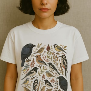 K&ouml;nnte beinhalten: Wei&szlig;es T-Shirt mit einer detaillierten Illustration verschiedener V&ouml;gel und Bl&auml;tter. Das Design zeigt eine Kr&auml;he, einen Specht und andere Vogelarten mit botanischen Elementen. Das Shirt hat einen klassischen Rundhalsausschnitt.