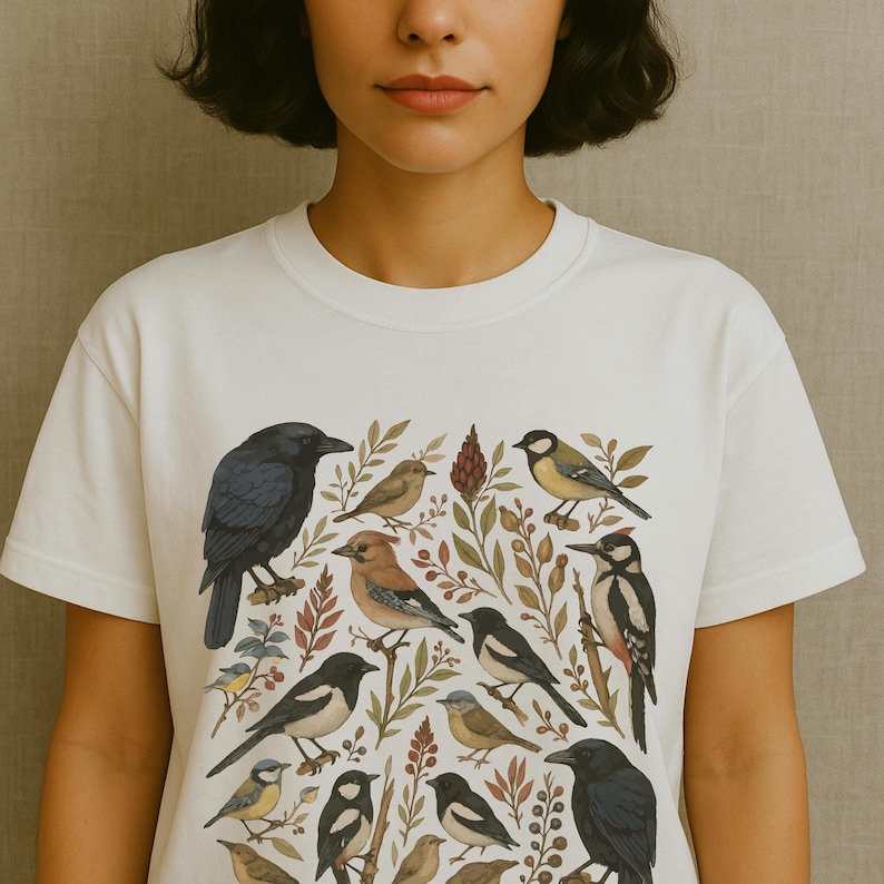 K&ouml;nnte beinhalten: Wei&szlig;es T-Shirt mit einer detaillierten Illustration verschiedener V&ouml;gel und Bl&auml;tter. Das Design zeigt eine Kr&auml;he, einen Specht und andere Vogelarten mit botanischen Elementen. Das Shirt hat einen klassischen Rundhalsausschnitt.