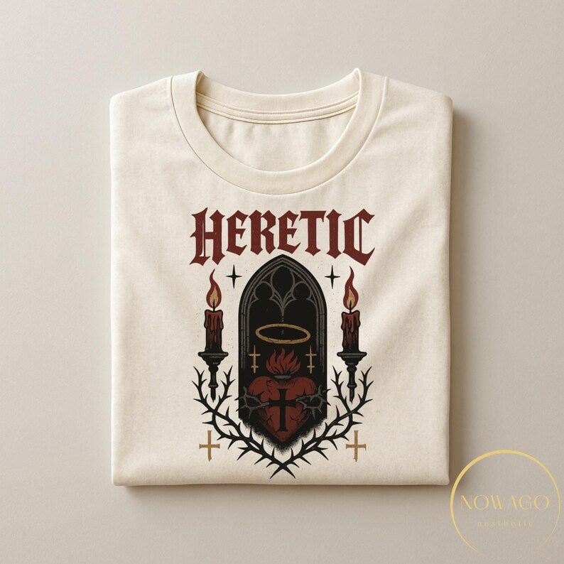 May include: Cream-colored t-shirt with the word "HERETIC" in a gothic font. The graphic features a heart with a cross, surrounded by thorns, candles, and a halo. The design is in shades of red, black, and cream.