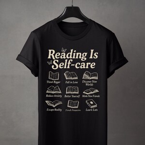 Puede incluir: Camiseta negra con el texto crema "Reading Is Self-care" e ilustraciones de libros. La camiseta incluye frases como "Think Bigger", "Fall in Love" y "Discover New Worlds".