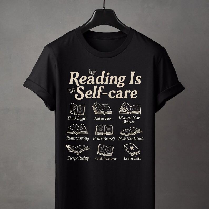 Puede incluir: Camiseta negra con el texto crema "Reading Is Self-care" e ilustraciones de libros. La camiseta incluye frases como "Think Bigger", "Fall in Love" y "Discover New Worlds".