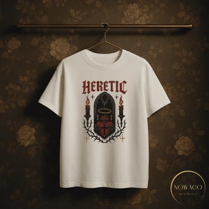 May include: White t-shirt with the word "HERETIC" in a gothic font above a graphic of a heart, candles, and a halo. The design is in red, black, and gold. The shirt is hanging on a metal bar.