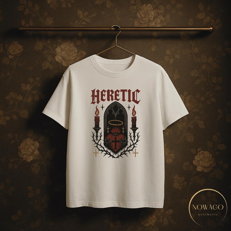 May include: White t-shirt with the word "HERETIC" in a gothic font above a graphic of a heart, candles, and a halo. The design is in red, black, and gold. The shirt is hanging on a metal bar.