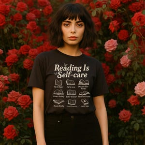 Puede incluir: Camiseta negra con el texto "Reading Is Self-care" e ilustraciones de libros. La camiseta enumera los beneficios de la lectura, como "Think Bigger" y "Discover New Worlds". El fondo presenta un rosal con rosas rojas y rosas.