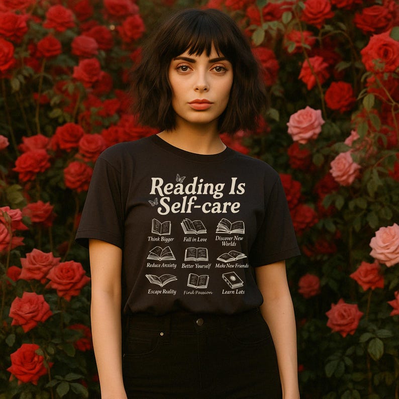 Puede incluir: Camiseta negra con el texto "Reading Is Self-care" e ilustraciones de libros. La camiseta enumera los beneficios de la lectura, como "Think Bigger" y "Discover New Worlds". El fondo presenta un rosal con rosas rojas y rosas.