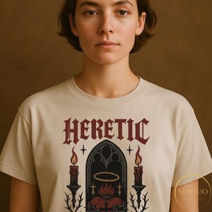 May include: Cream-colored t-shirt featuring the word "HERETIC" in a gothic font above a graphic of a church window, candles, and a heart. The design is in shades of red, black, and gold. The shirt has a crew neck and short sleeves.