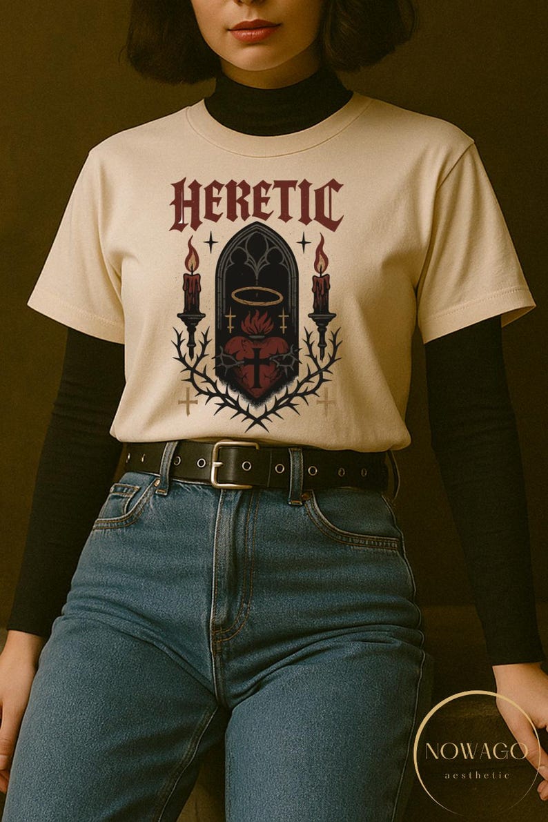 May include: Cream-colored t-shirt with the word "HERETIC" in burgundy gothic lettering. The shirt features a central graphic of a heart, cross, and thorns within a gothic arch, flanked by candles. The model is wearing blue jeans and a black belt.