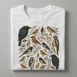 K&ouml;nnte beinhalten: Wei&szlig;es T-Shirt mit einer detaillierten Illustration verschiedener V&ouml;gel, darunter eine Kr&auml;he, ein Specht und eine Elster, inmitten botanischer Elemente. Das Design integriert eine Reihe von Farben, von Schwarz und Braun bis hin zu Gelb und Gr&uuml;n, wodurch eine von der Natur inspirierte &Auml;sthetik entsteht.