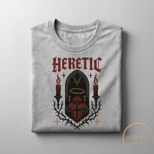 May include: A light gray crew neck t-shirt featuring a gothic design. The design includes the word "HERETIC" in a gothic font, a heart with a cross, candles, and a halo. The graphic is in shades of red, black, and gold.