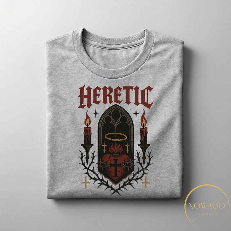 May include: A light gray crew neck t-shirt featuring a gothic design. The design includes the word "HERETIC" in a gothic font, a heart with a cross, candles, and a halo. The graphic is in shades of red, black, and gold.