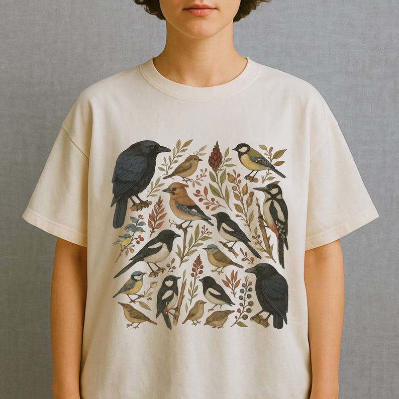 K&ouml;nnte beinhalten: Cremefarbenes T-Shirt mit einer detaillierten Illustration verschiedener Vogelarten, darunter Kr&auml;hen, Eichelh&auml;her und Spechte, inmitten botanischer Elemente. Das Design zeigt eine Reihe von Farben, von tiefem Schwarz und Blau bis hin zu erdigem Braun und Gr&uuml;n.