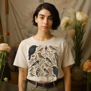 K&ouml;nnte beinhalten: Cremefarbenes T-Shirt mit einer detaillierten Illustration verschiedener V&ouml;gel und Bl&auml;tter. Das Design zeigt V&ouml;gel in Schwarz, Braun, Gelb und Wei&szlig; vor dem Hintergrund gr&uuml;ner und brauner Bl&auml;tter. Das Shirt hat einen klassischen Rundhalsausschnitt und kurze &Auml;rmel.