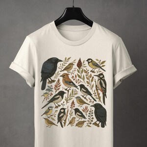 K&ouml;nnte beinhalten: Cremefarbenes T-Shirt mit einer detaillierten Illustration verschiedener V&ouml;gel, darunter Kr&auml;hen, Elstern und Spechte, inmitten botanischer Elemente. Das Design zeigt eine Vielzahl von Vogelarten in einem naturalistischen Stil, mit Erdt&ouml;nen und komplizierten Details.