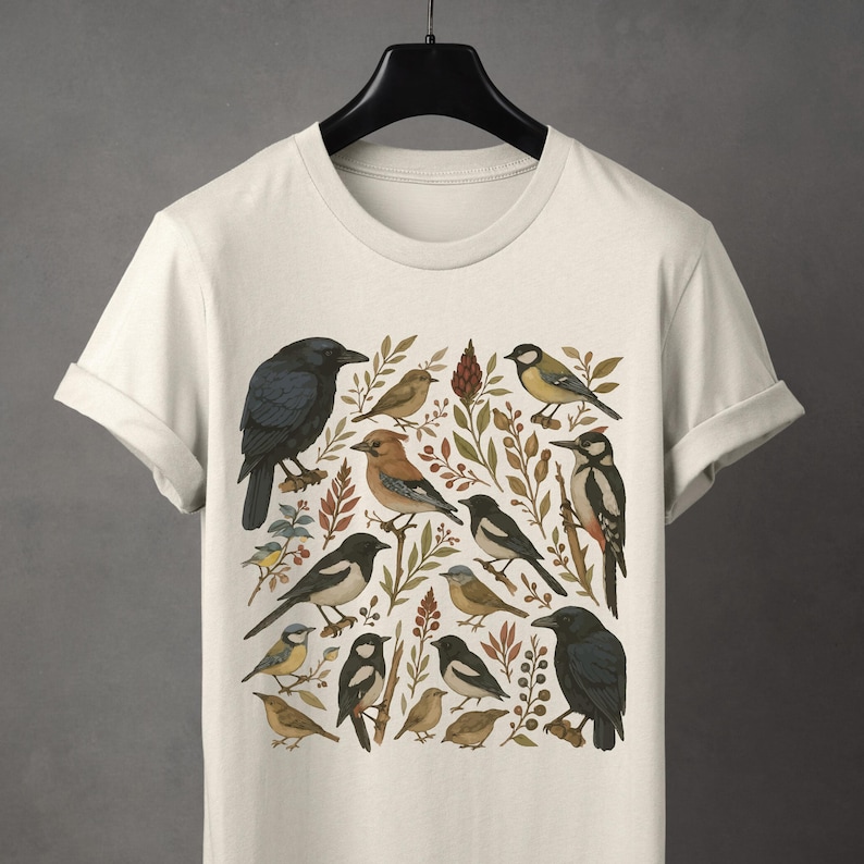 K&ouml;nnte beinhalten: Cremefarbenes T-Shirt mit einer detaillierten Illustration verschiedener V&ouml;gel, darunter Kr&auml;hen, Elstern und Spechte, inmitten botanischer Elemente. Das Design zeigt eine Vielzahl von Vogelarten in einem naturalistischen Stil, mit Erdt&ouml;nen und komplizierten Details.