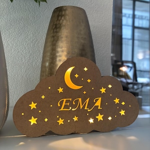 Peut inclure: Veilleuse en forme de nuage en bois marron avec le nom "EMA" en lettres dorées. Le design comprend un croissant de lune et des étoiles découpées qui émettent une lueur chaude. La veilleuse est sur une surface blanche.