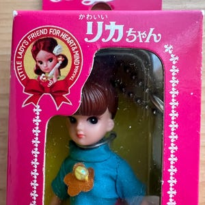 May include: A vintage Licca doll in its original packaging. The doll has brown hair and is wearing a blue dress with a flower accent. The box is pink with Japanese text and the words "Little Lady's Friend for Heart & Mind".