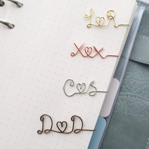 Könnte beinhalten: Vier Draht-Lesezeichen in Gold, Kupfer, Silber und Bronze. Jedes Lesezeichen hat ein einzigartiges Design: "L♡R", "X♡X", "C♡S" und "D♡D". Die Lesezeichen befinden sich auf einer weißen, gepunkteten Notizbuchseite.