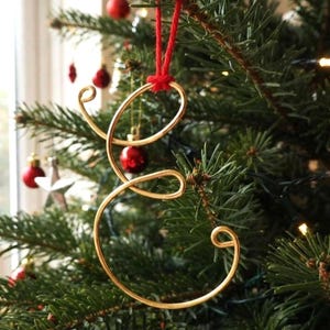 May include: A gold-coloured wire ornament shaped like the cursive letter E. The ornament features smooth, continuous loops and curves, and it is suspended by a bright red string tied at the top, creating a decorative hanging piece for display.
