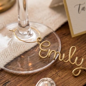 Peut inclure: Un marque-verre en métal doré présentant le prénom Emily écrit en lettres cursives continues en fil de fer. Le marque-verre est fixé à un anneau circulaire conçu pour s'adapter autour du pied d'un verre, avec une finition métallique.