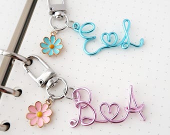 May include: Two metal keychains featuring silver-tone lobster clasps. One keychain has a light blue wire script spelling EL with a heart and a light blue daisy charm. The other has a pink wire script spelling BA with a heart and a pink daisy charm.