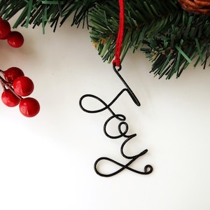 Puede incluir: Un adorno de alambre negro con la palabra Joy en letra cursiva, suspendido por un hilo rojo fino. El adorno se sitúa cerca de agujas de pino verdes y un racimo de pequeñas bayas rojas brillantes, formando una decoración festiva simple.