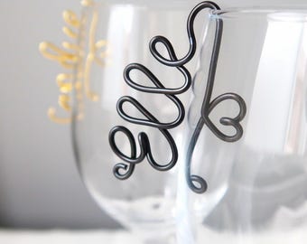 May include: A black wire wine glass charm shaped into the cursive word "elle" with a small heart detail attached to the rim of a clear glass. A second gold wire charm is partially visible on a separate glass in the background.