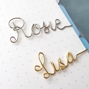 May include: Two wire name bookmarks. One is silver and spells "Rosie", the other is gold and spells "Lisa". Both are in a cursive font. The background is a white notebook page with small grey dots.