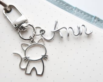 May include: A silver-toned metal keychain featuring a lobster clasp, a cat-shaped outline charm, and a wire-formed name charm that reads Ann. The keychain components are connected by small circular jump rings, creating a dangling accessory design.
