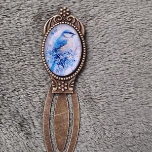 Puede incluir: Marcapáginas de metal color bronce con una imagen ovalada de un pájaro azul posado en una rama con flores azules. El marcapáginas tiene una parte superior decorativa y un cuerpo largo y delgado.