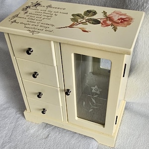 May include: A cream-coloured wooden jewellery cabinet featuring four small drawers with dark knobs and a side door with a glass panel etched with a flower design. The top surface displays a pink rose illustration and the text Pink Rosebud.