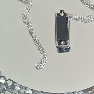 May include: A silver chain necklace with a black harmonica pendant. The necklace is lying on a white surface with a border of rhinestones.